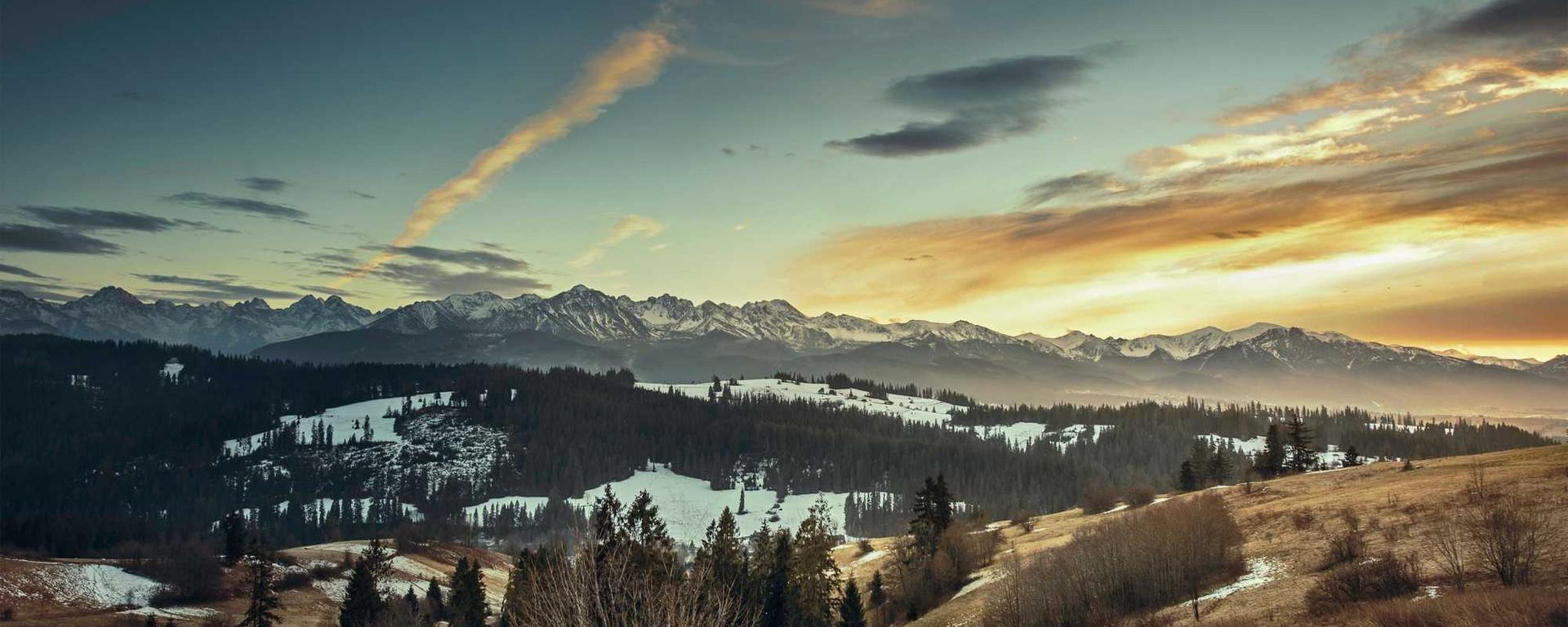 Landscape photo of wooded hills with patches of snow, and mountains in the distance.